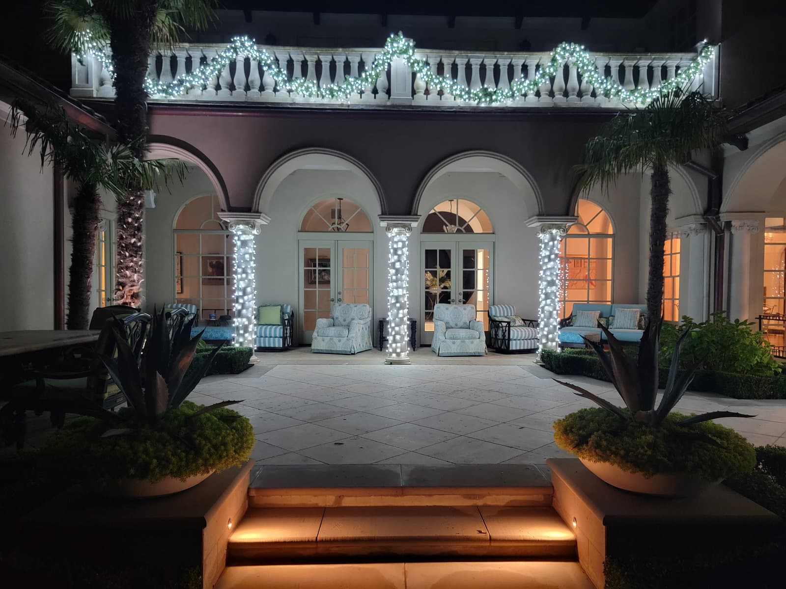 Courtyard columns wrapped in cool white mini lights beneath a balcony draped in lighted garland