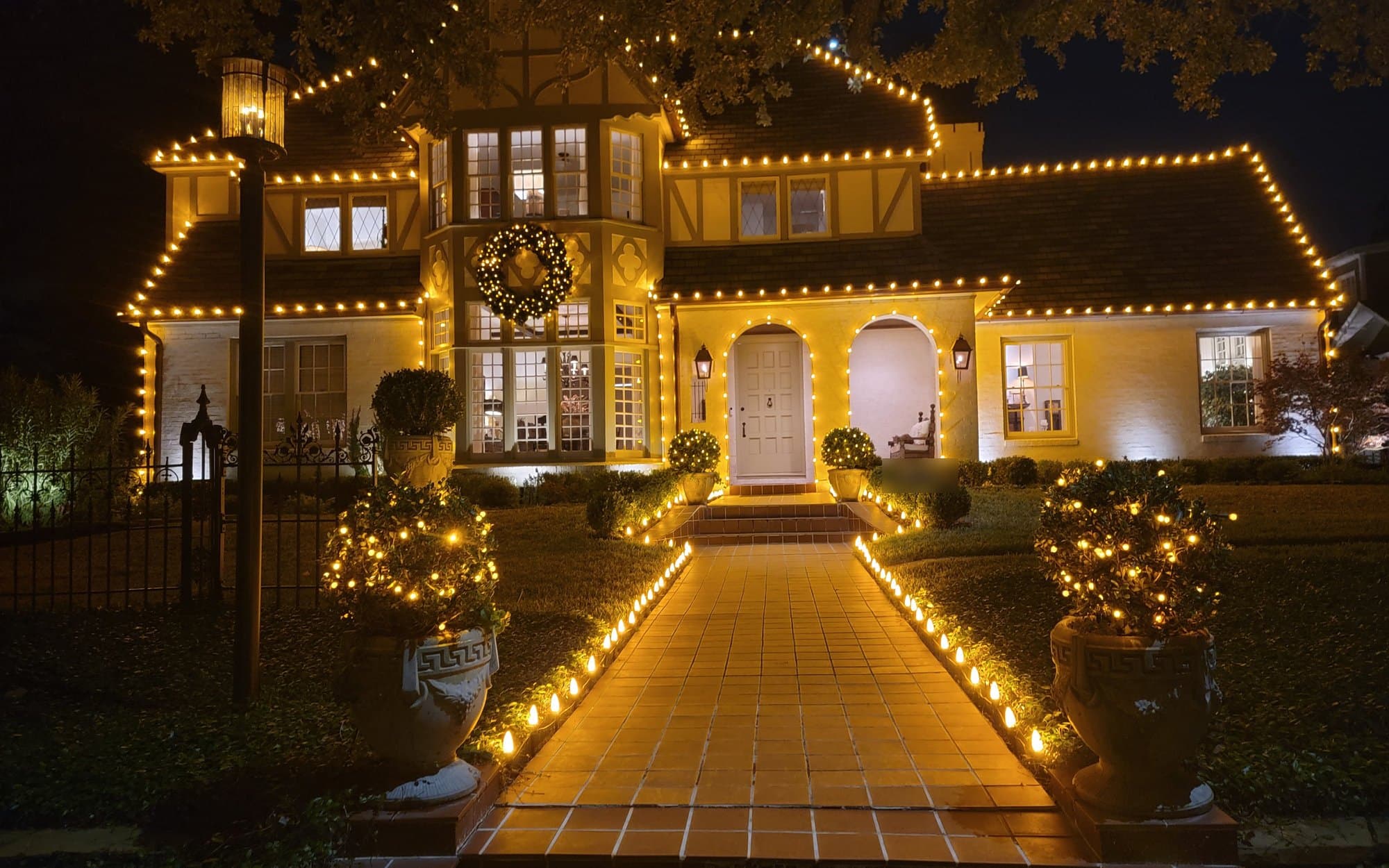 Tudor-style home outlined in warm white C9 lights, with a lit wreath, wrapped topiaries and a lighted front walkway